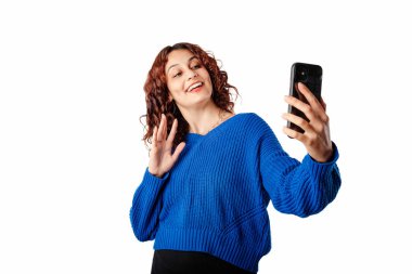 Cheerful woman wearing blue pullover sweater isolated over white background making a video call with smartphone and waving palm hand. Talks remotely with boyfriend or family. Laughs. Doing selfie.