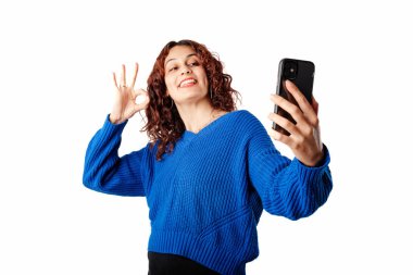 Happy woman wearing sweater standing isolated over white background taking a selfie with the okay sign, looks at the phone and smiles. Video call with family. Video call with hearing impaired friend.