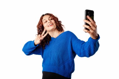 Young girl smiling happy wearing pullover sweater isolated over white background makes a video call or selfie with smartphone makes a peace symbol with fingers. Strikes a friendly pose. Number two.
