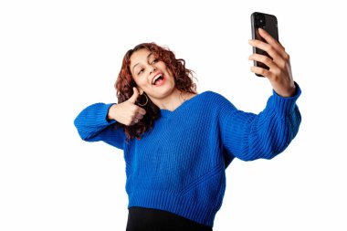 Young woman smiling wearing sweater standing isolated over white background taking selfie and gesturing like, thumbs up to smartphone, communicating by video call. I approve, I agree with you.