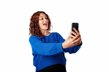 Portrait of woman wearing blue knitted sweater standing isolated over white background excited, looks at the phone screen with a happy expression. Overjoyed.