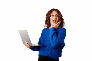 Portrait of woman wearing blue knitted sweater standing isolated over white background shocked, extremely surprised, holding a laptop touches face with palm and looks at the camera.