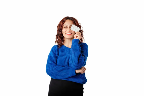 Happy woman wearing pullover sweater standing isolated over white background cover one eye with holding a credit card in hand, laughs, looks at the camera and poses. Shopping concept.