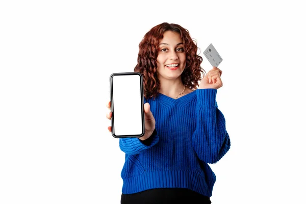 Young woman smiling confident standing isolated over white background shows the camera blank white mockup screen of the phone and holds a credit card in the other hand. Smiles looking at the camera.