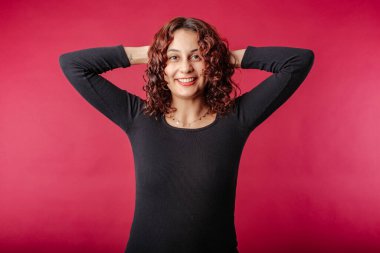 Happy woman wearing black dress standing isolated over red background relaxing and stretching, arms and hands behind head and neck. Success concept.