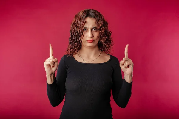Beautiful redhead woman wearing black dress standing isolated over red background pointing up, furrows, frowning eyebrows upset, need explanation. Feel unfair or displeased, complaining.