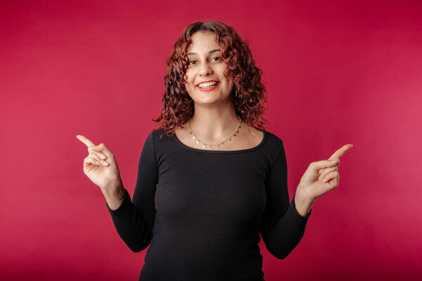 Happy woman wearing black dress standing isolated over red background with a happy expression, pointing left and right with index fingers. Pointing to opposite sides.