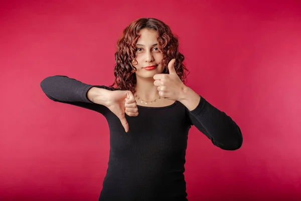 Portrait of cheerful woman wearing black ribbed dress standing isolated over red background doing thumbs up and down, disagreement and agreement expression. She looks unsure.