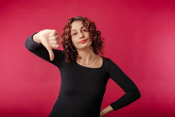 Young redhead woman wearing black ribbed dress isolated over red background not believing the lies, looks at the camera and gives the thumbs down sign. Don\'t tell lies. I didn\'t believe it.
