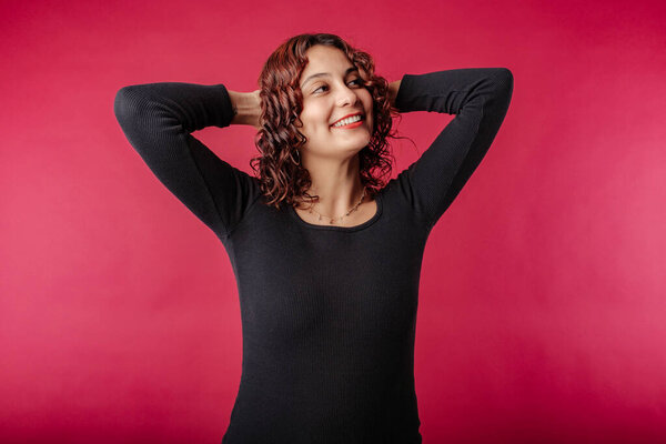 Young redhead girl smiling happy wearing black ribbed dress isolated over red background relaxing and stretching, arms and hands behind head and neck. Success concept.