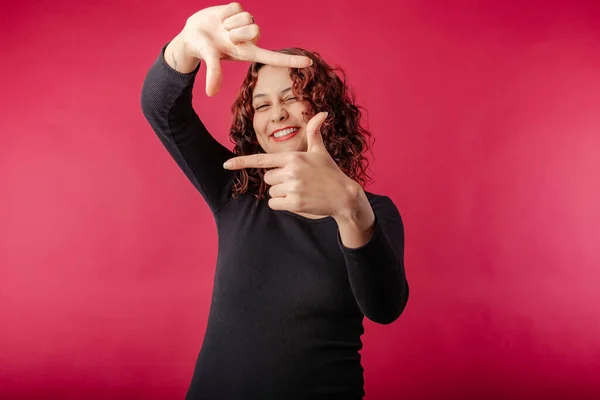 Young redhead girl smiling happy wearing black ribbed dress isolated over red background perfectly playful, doing frame gesture with hands and smiling. Frame, recording, archive, camera, movie concept