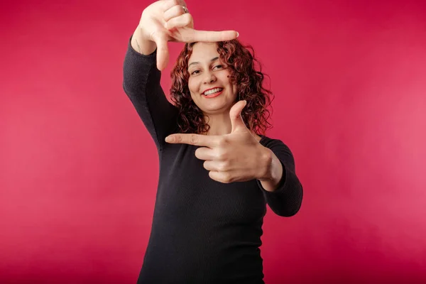 Cute caucasian woman wearing black ribbed dress isolated over red background perfectly playful, doing frame gesture with hands and smiling. Frame, recording, archive, camera, movie concept.