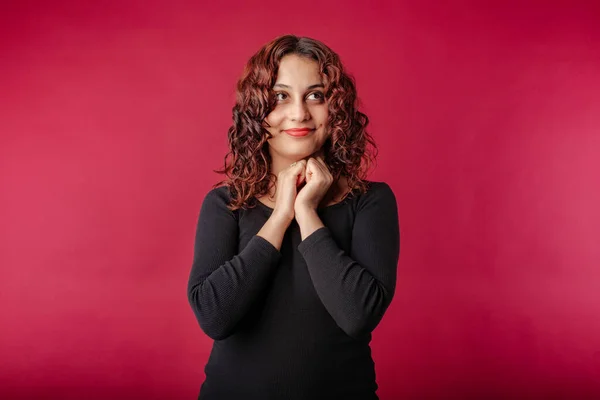 Redhead millennial woman wearing black dress standing isolated over red background imagining and thinking with her hands together. Looking to empty corner copy space.