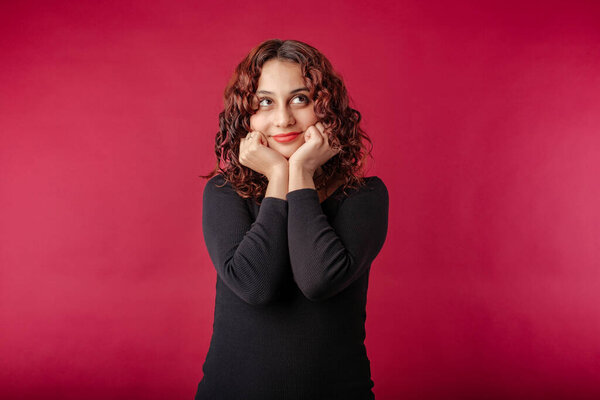 Cute redhead woman wearing black dress standing isolated over red background touching chin with fists, looks at the corner of the empty copy space with hopeful eyes.
