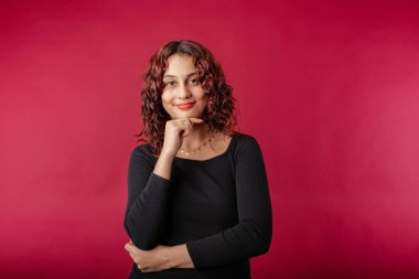 Beautiful redhead woman wearing black dress standing isolated over red background touches chin and looks suspiciously at the camera. Trying to find ideas.