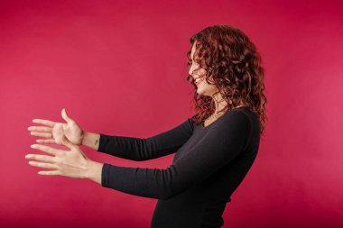 Side view of young redhead woman standing isolated over red background outstretching hands as if giving for free, offering to embrace, complacency and egoism concept. Satisfy longing, share excitement
