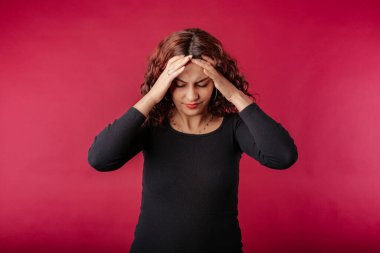 Portrait of cute redhead woman wearing black ribbed dress standing isolated over red background holding head in discomfort due to pain. Headache.