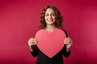 Cute caucasian woman wearing black ribbed dress isolated over red background holding a large cardboard heart shape smiles at the camera. The concept of love, intimacy, togetherness. Looks with love.