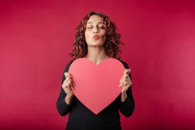 Cute caucasian woman wearing black ribbed dress isolated over red background puckering lips in a kiss, poses with a large cardboard heart shape in hand. Eyes are closed, thinking of lover.