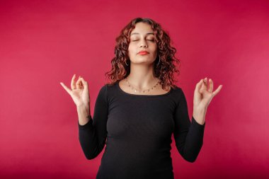 Woman standing isolated over red background keeping eyes closed, holding fingers in mudra gesture. Meditation, religion and spiritual practices. Peace and calm. Releases all tension and stress.