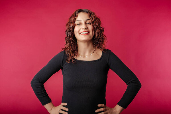 Beautiful redhead woman wearing black dress standing isolated over red background enjoys talking and laughs a lot. He touches his chin with his hand and likes it.