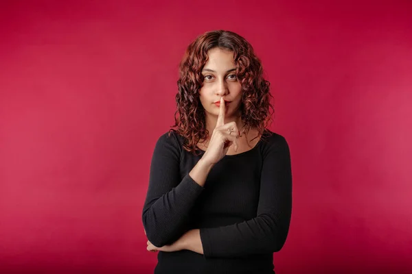 Young beautiful woman wearing black ribbed dress isolated over red background asking to be quiet with finger on lips. Silence and secret seriousness concept.