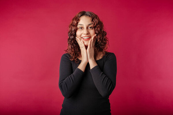 Young redhead girl smiling happy wearing black ribbed dress isolated over red background staring amazed touching cheek. Looks at the camera very excitedly.