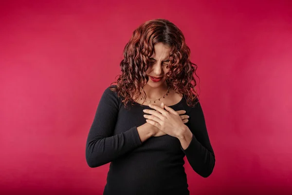 Young beautiful woman wearing black ribbed dress isolated over red background having severe chest pain as heart attack and illness concept. Hold hand on chest heart crying posing.