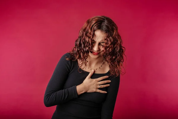 Redhead millennial woman wearing black dress standing isolated over red background having severe chest pain as heart attack and illness concept. Hold hand on chest heart crying posing.