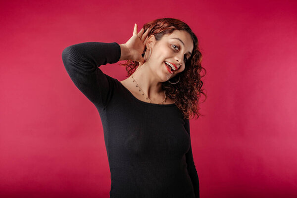 Young woman smiling confident wearing black dress standing isolated over red background smiling with hand over ear listening an hearing to rumor or gossip. Deafness concept. All ears and very curious.