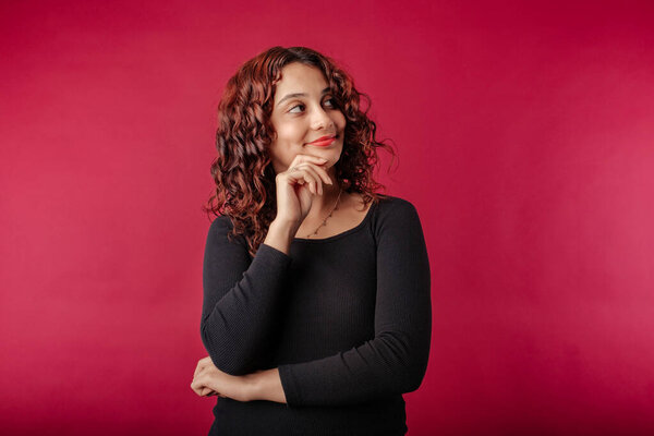 Young redhead girl happy isolated over red background touching cheek with index finger, laughs and looks at the empty copy corner. Thoughtful and decided what wants. Looks at the advertising space.