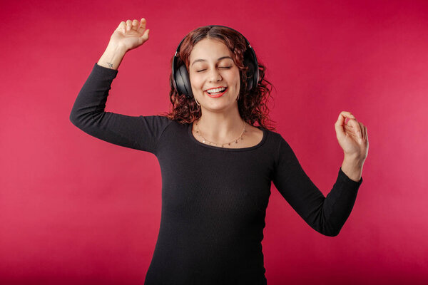 Happy redhead woman wearing black ribbed dress standing isolated over red background listen music with headphones, dancing. Dancing to favorite music is very happy. Enjoys it very much.