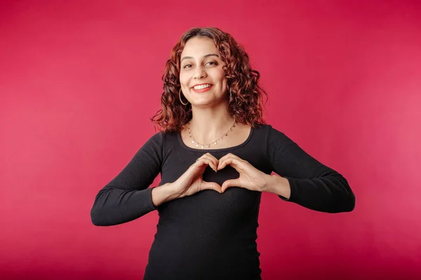Cute redhead woman wearing black dress standing isolated over red background making a heart gesture with her fingers in front of her chest showing her love. With love, you are not alone. I love you.