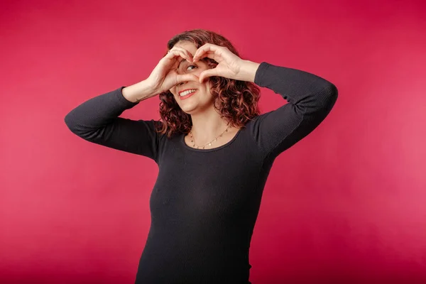 Happy redhead woman standing isolated over red background doing heart shape with hand and fingers looking through sign. Smiles with love. Makes a heart sign with hands and looks at the camera, at you.