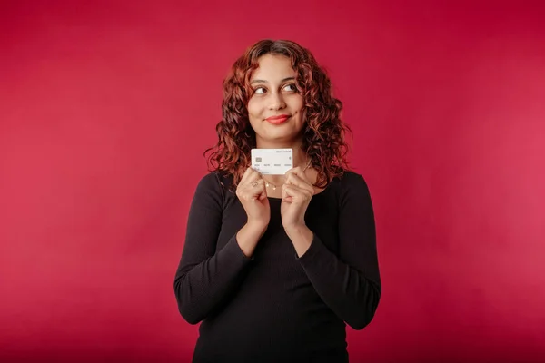 Young redhead woman wearing ribbed dress isolated over red background holding a credit card looks at the empty copy space and daydreams. Deciding what to buy online. Thinking about sales and purchases