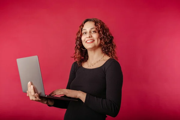 Young woman smiling confident standing isolated over red background holding a laptop, smiling at the camera with a confident expression. Typing on laptop keyboard. Creating web design or digital work.