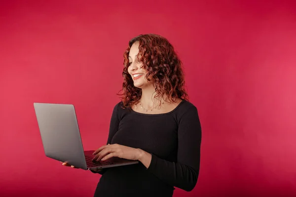 Young woman confident standing isolated over red background holding a laptop with a confident expression. Typing on laptop keyboard. Creating web design or digital work. Looking at the computer screen
