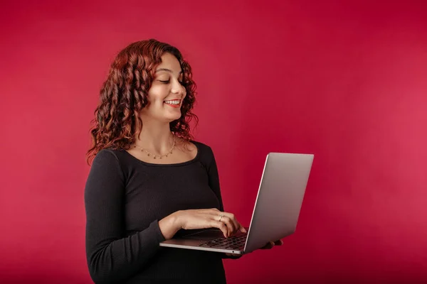 Young woman confident standing isolated over red background holding a laptop with a confident expression. Typing on laptop keyboard. Creating web design or digital work. Looking at the computer screen