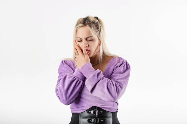 Young blonde woman wearing lilac ribbed blouse isolated over white background with mouth open, touching cheek with both hands, is suffering from a toothache. Looks very sad.