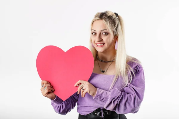 Young woman smiling confident wearing ribbed blouse standing isolated over white background poses smiling with a cardboard heart shape. Looking at the camera, at you, at lover. It could be a gift.