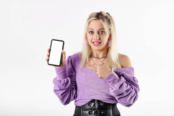 Blonde woman standing isolated over white background excited, showing phone pointing finger at blank white screen. Mobile app. An incredible campaign or discount news. Showing online application.