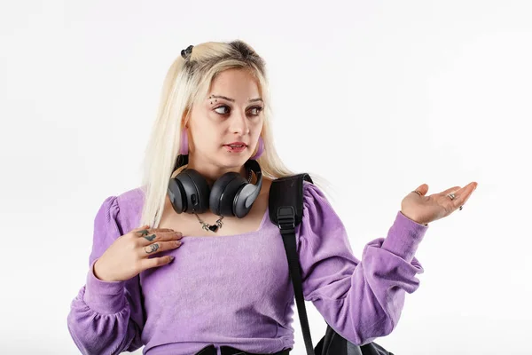 Blonde millennial woman wearing ribbed blouse standing isolated over white background with headphones around neck, offering empty copy space with palm hand.