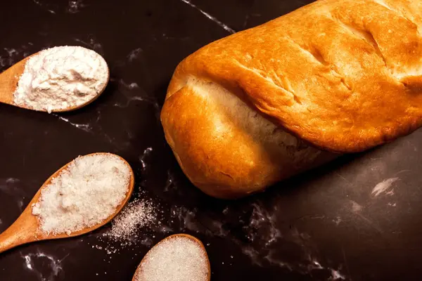 新鮮な自家製パンと黒い大理石の背景に砂糖塩と小麦粉の3つの木製のスプーン — ストック写真
