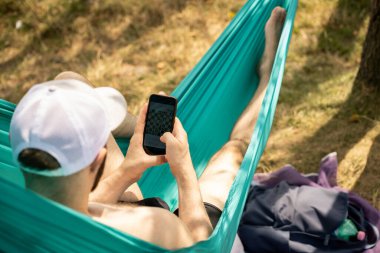 Bir adam hamakta dinlenirken telefonda satranç oynuyor.