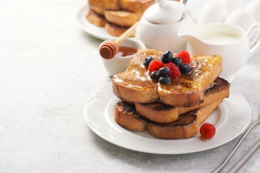 Yumurtaya, süte ve şekere batırılmış, tavada kızartılmış, beyaz tabakta taze ahududu, yaban mersini, bal, açık gri zemin üzerine nane içeren birkaç parça Fransız tostu.