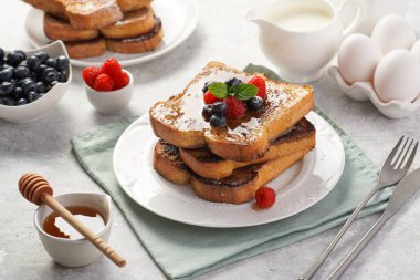 Yumurtaya, süte ve şekere batırılmış, tavada kızartılmış, beyaz tabakta taze ahududu, yaban mersini, bal, açık gri zemin üzerine nane içeren birkaç parça Fransız tostu.