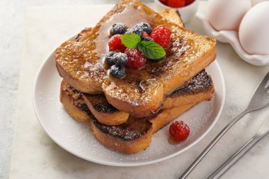 Yumurtaya, süte ve şekere batırılmış, tavada kızartılmış, beyaz tabakta taze ahududu, yaban mersini, bal, açık gri zemin üzerine nane içeren birkaç parça Fransız tostu.
