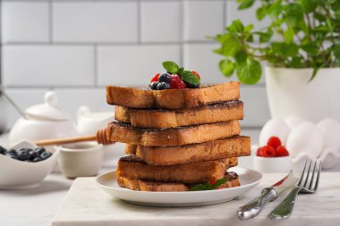 Yumurtaya, süte ve şekere batırılmış, tavada kızartılmış, beyaz tabakta taze ahududu, yaban mersini, bal, açık gri zemin üzerine nane içeren birkaç parça Fransız tostu.