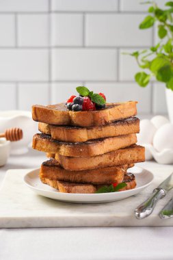 Yumurtaya, süte ve şekere batırılmış, tavada kızartılmış, beyaz tabakta taze ahududu, yaban mersini, bal, açık gri zemin üzerine nane içeren birkaç parça Fransız tostu.