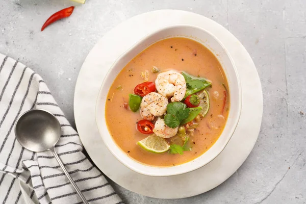 a spicy thai coconut soup or curry made of coconut milk, cherry tomatoes, snow pea, chili pepper, lime, ginger, shrimps and fresh cilantro on marble board on grey concrete background, top view